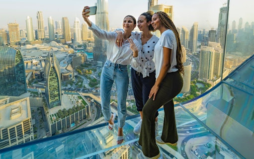 Visitors taking a selfie on the glass floor of Sky Views Observatory, Dubai skyline in background.