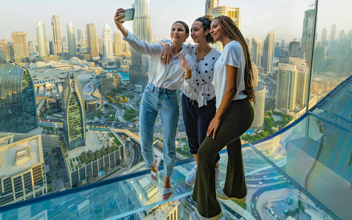 Visitors taking a selfie on the glass floor of Sky Views Observatory, Dubai skyline in background.