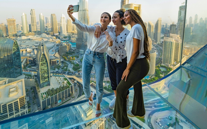 Visitors taking a selfie on the glass floor of Sky Views Observatory, Dubai skyline in background.