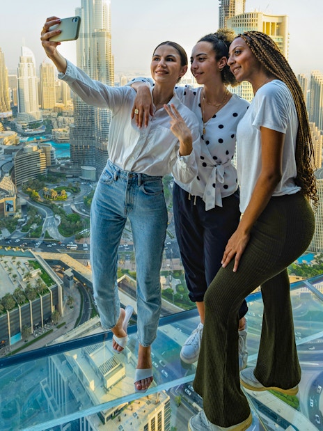 Visitors taking a selfie on the glass floor of Sky Views Observatory, Dubai skyline in background.