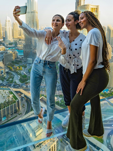 Visitors taking a selfie on the glass floor of Sky Views Observatory, Dubai skyline in background.