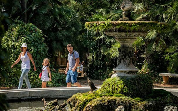 Family walking by a fountain in Parc Samà, surrounded by lush greenery.