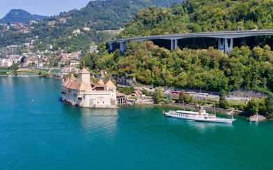 Cruise boat approaching Chillon Castle on Lake Geneva near Vevey, surrounded by lush hills.