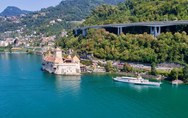 Cruise boat approaching Chillon Castle on Lake Geneva near Vevey, surrounded by lush hills.