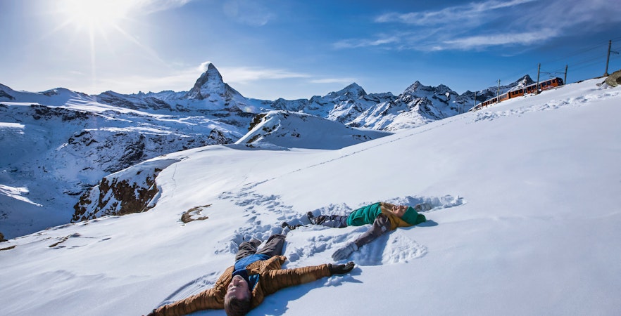People making snow angels with Matterhorn in the background, Zermatt to Gornergrat train visible.