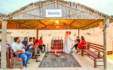 Shisha lounge in desert safari with people enjoying hookah under a thatched roof.