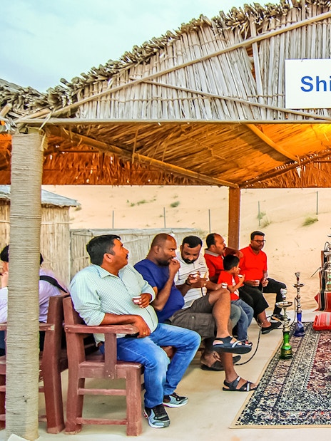 Shisha lounge in desert safari with people enjoying hookah under a thatched roof.