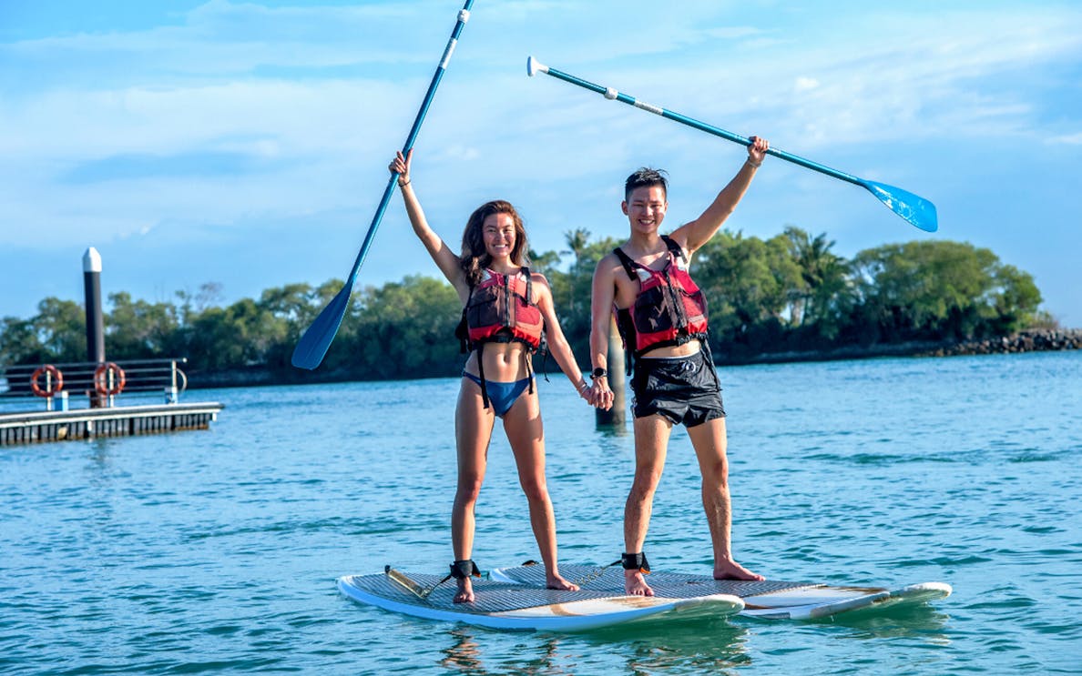 Couple paddleboarding at Ola Beach Club, Singapore, holding paddles aloft.