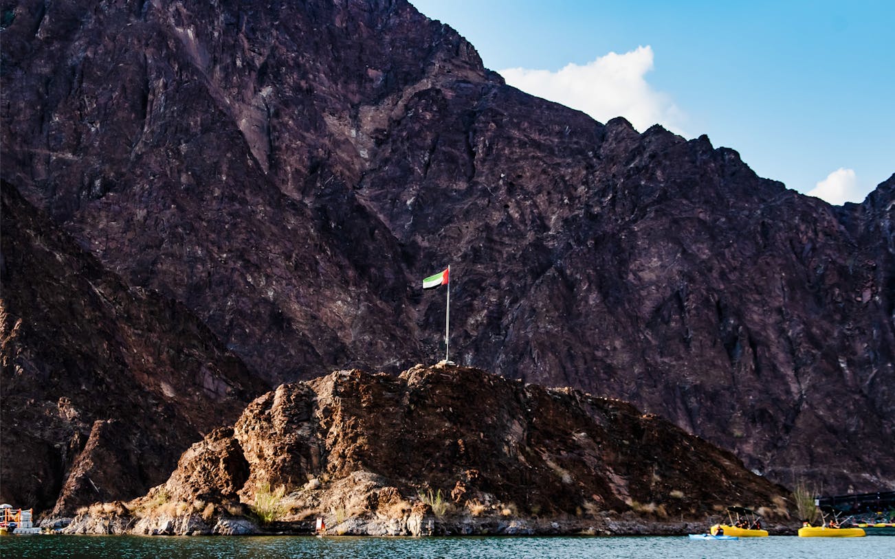 Hatta Dam with UAE flag on rocky hill, surrounded by mountains and water.