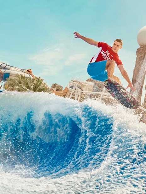 Surfer riding wave at Yas Waterworld Abu Dhabi with water slides in background.
