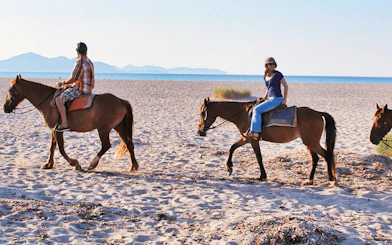 Horseback riders on a sandy beach in Mallorca with distant mountains.