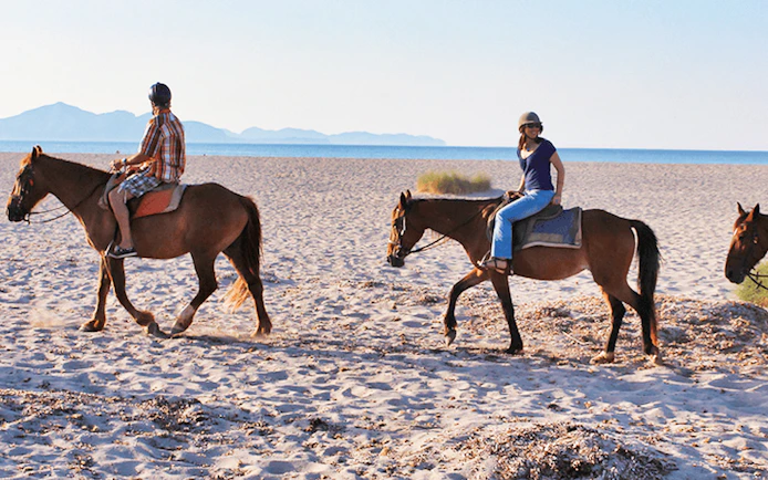 Horseback riders on a sandy beach in Mallorca with distant mountains.