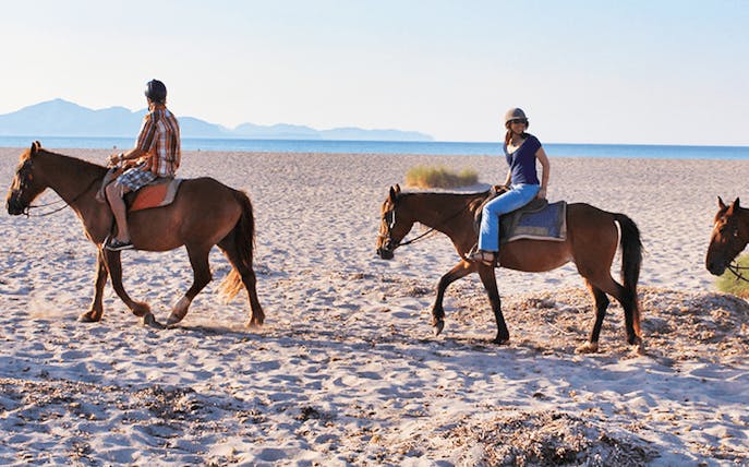 Horseback riders on a sandy beach in Mallorca with distant mountains.