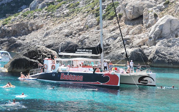 Robinson catamaran with tourists in Pollensa Bay, Mallorca, near rocky coastline.