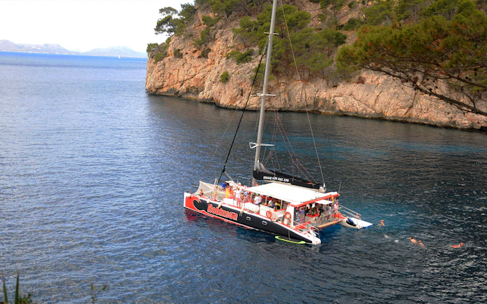 Robinson catamaran with swimmers in Pollensa Bay near rocky cliffs.