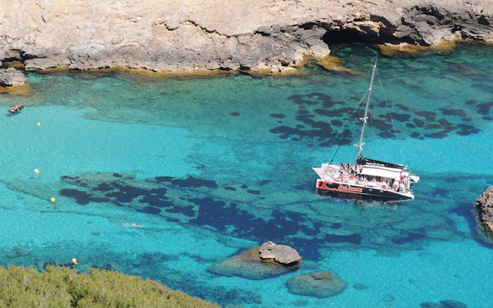 Catamaran sailing in clear waters of Pollensa Bay, Mallorca, with rocky coastline.