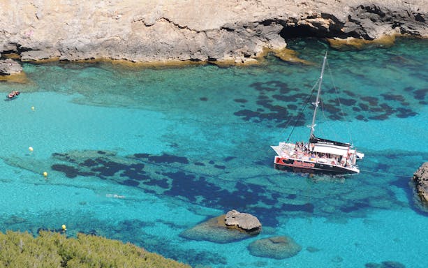 Catamaran sailing in clear waters of Pollensa Bay, Mallorca, with rocky coastline.