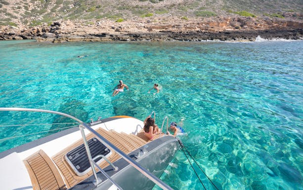 Catamaran anchored near rocky shore with people swimming in clear waters, Palma de Mallorca.