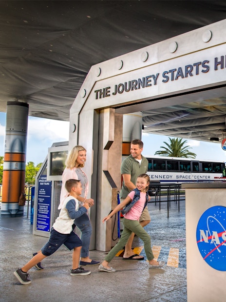 Family entering Kennedy Space Center with NASA logo and tour bus in background.