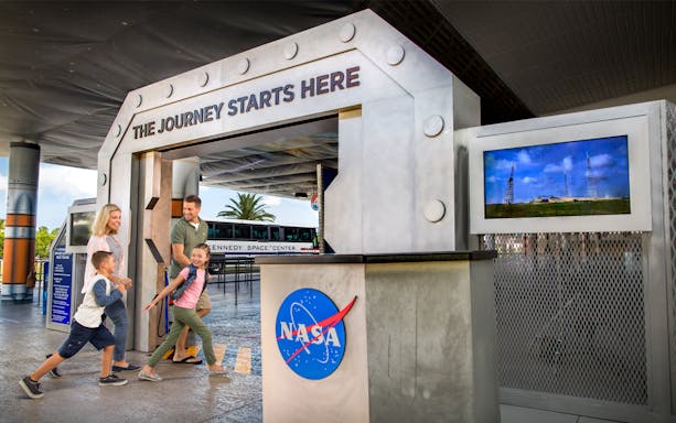 Family entering Kennedy Space Center with NASA logo and tour bus in background.