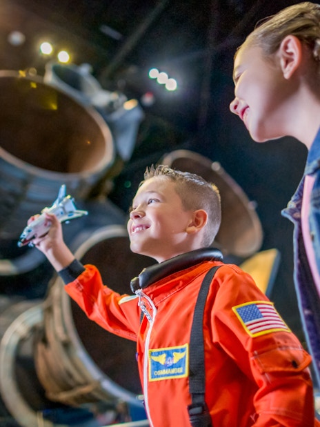 Child in astronaut suit holding model shuttle at Kennedy Space Center exhibit.