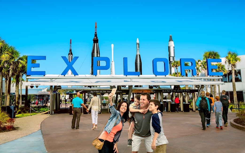 Family taking a selfie at the entrance of Kennedy Space Center with rockets in the background.