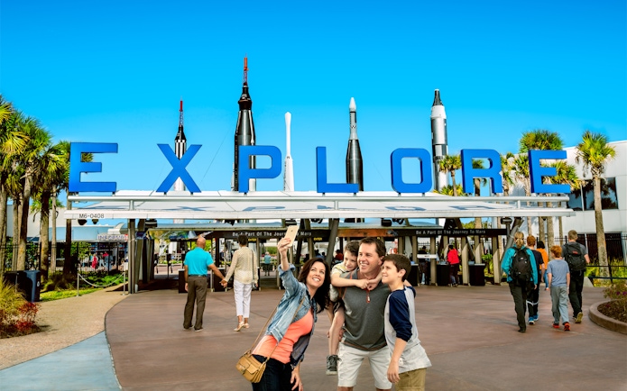 Family taking a selfie at the entrance of Kennedy Space Center with rockets in the background.