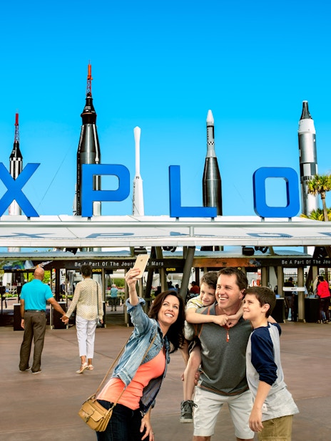 Family taking a selfie at the entrance of Kennedy Space Center with rockets in the background.