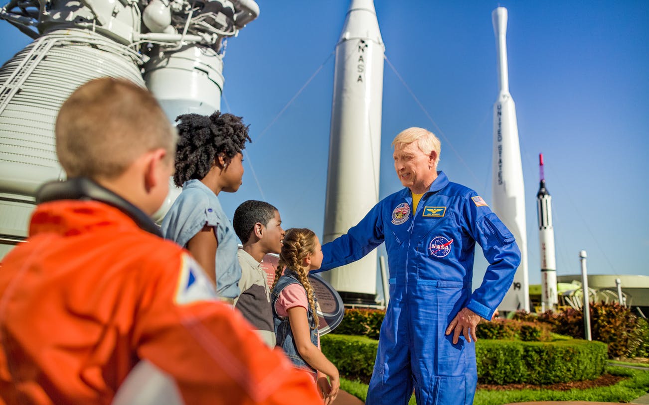 Children learning from a guide in a NASA suit at Kennedy Space Centre with rockets in the background.