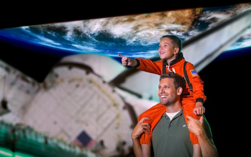 Child in astronaut suit on adult's shoulders at Kennedy Space Centre, Earth backdrop.