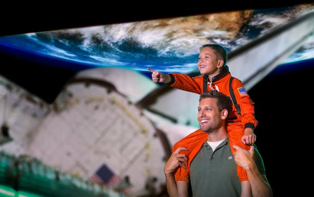 Child in astronaut suit on adult's shoulders at Kennedy Space Centre, Earth backdrop.