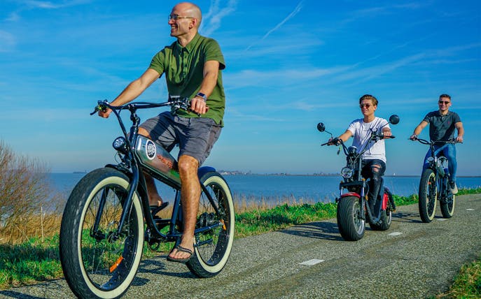 Cyclists riding e-fatbikes along a scenic path by the water in Volendam, Amsterdam countryside.
