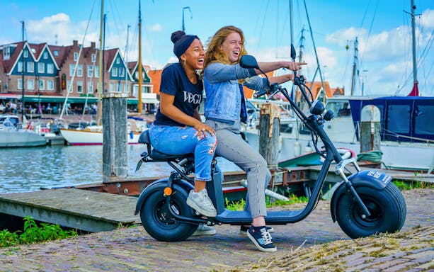 Two people on an e-scooter by the harbor in Volendam, Amsterdam countryside.