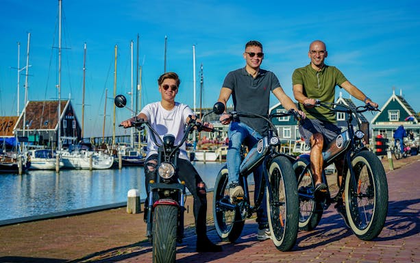 Three people riding e-scooters along a marina in Volendam, Amsterdam countryside.