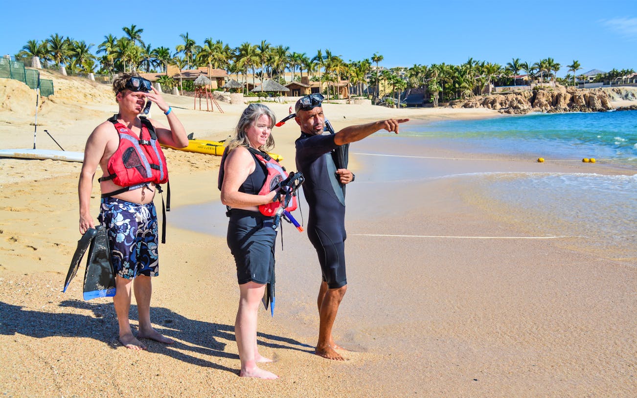 Guide pointing out snorkeling spots to tourists on a sandy beach in Cabo San Lucas.