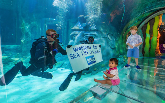 Diver holding "Welcome to SEA LIFE Orlando" sign in aquarium with children watching.