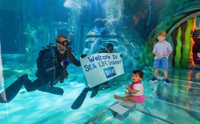 Diver holding "Welcome to SEA LIFE Orlando" sign in aquarium with children watching.