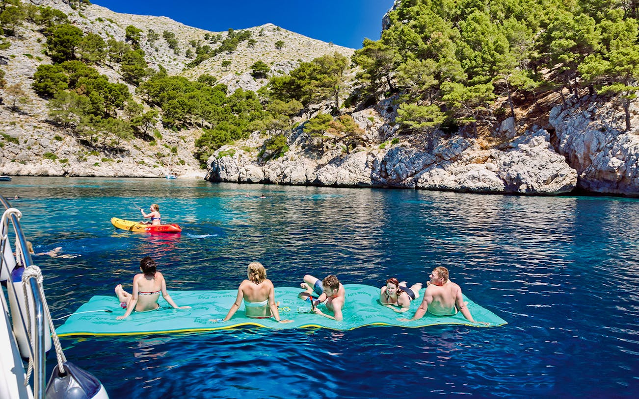 Small group enjoying a boat tour in Alcudia, relaxing on a floating mat with scenic rocky coastline.