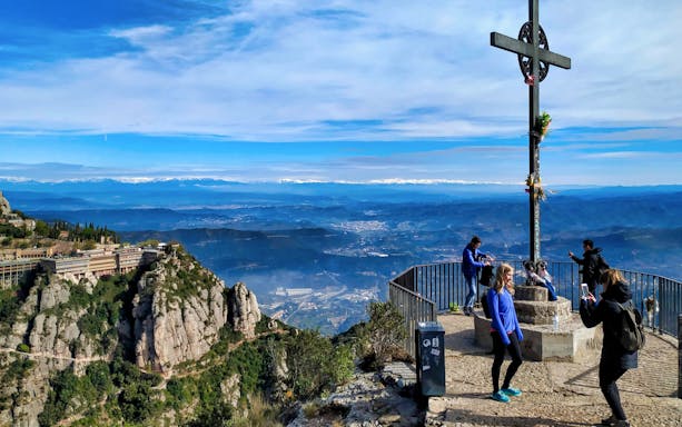 Hikers at Montserrat Monastery viewpoint with cross, overlooking Catalonia landscape.