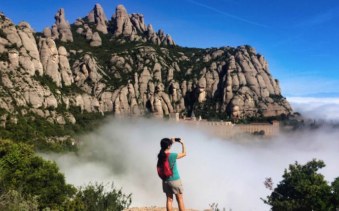 Hiker photographing Montserrat Monastery surrounded by misty mountains, Spain.