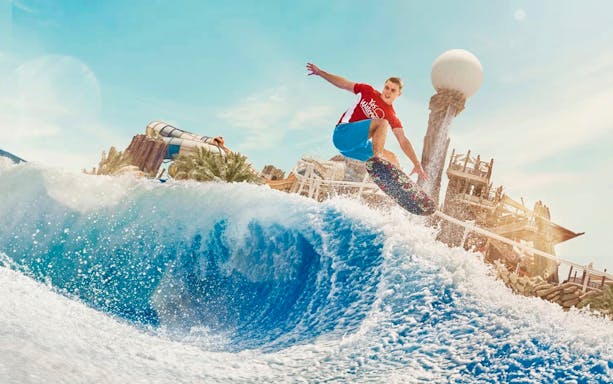 Surfer riding wave at Yas Waterworld Abu Dhabi with water slides in background.