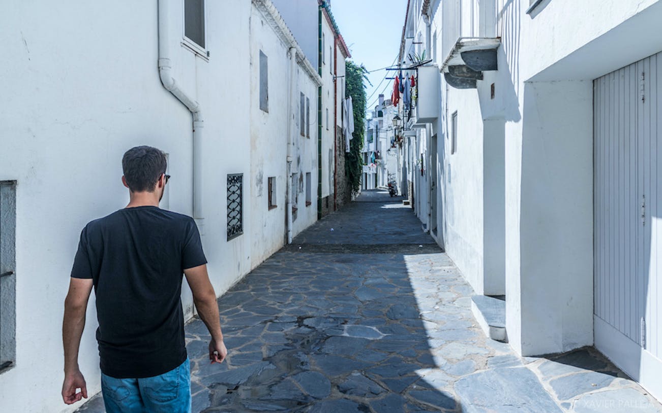 Man walking down a narrow street in Cadaqués, Spain, with whitewashed buildings.