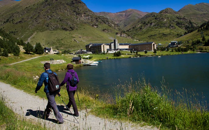 Hikers walking along a lakeside trail in Vall de Núria, Pyrenees, with mountains in the background.
