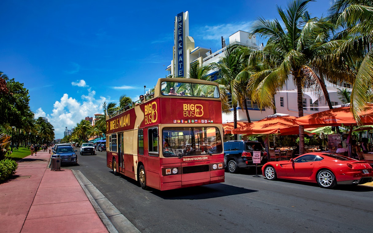 Open-top tour bus on Ocean Drive, Miami, with palm trees and art deco buildings.