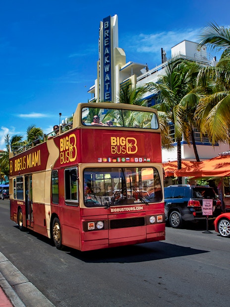 Open-top tour bus on Ocean Drive, Miami, with palm trees and art deco buildings.