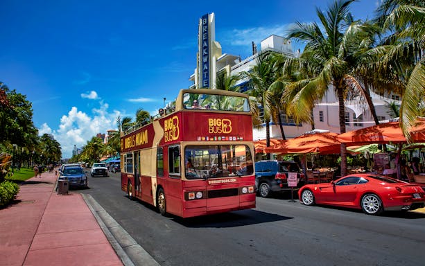 Open-top tour bus on Ocean Drive, Miami, with palm trees and art deco buildings.