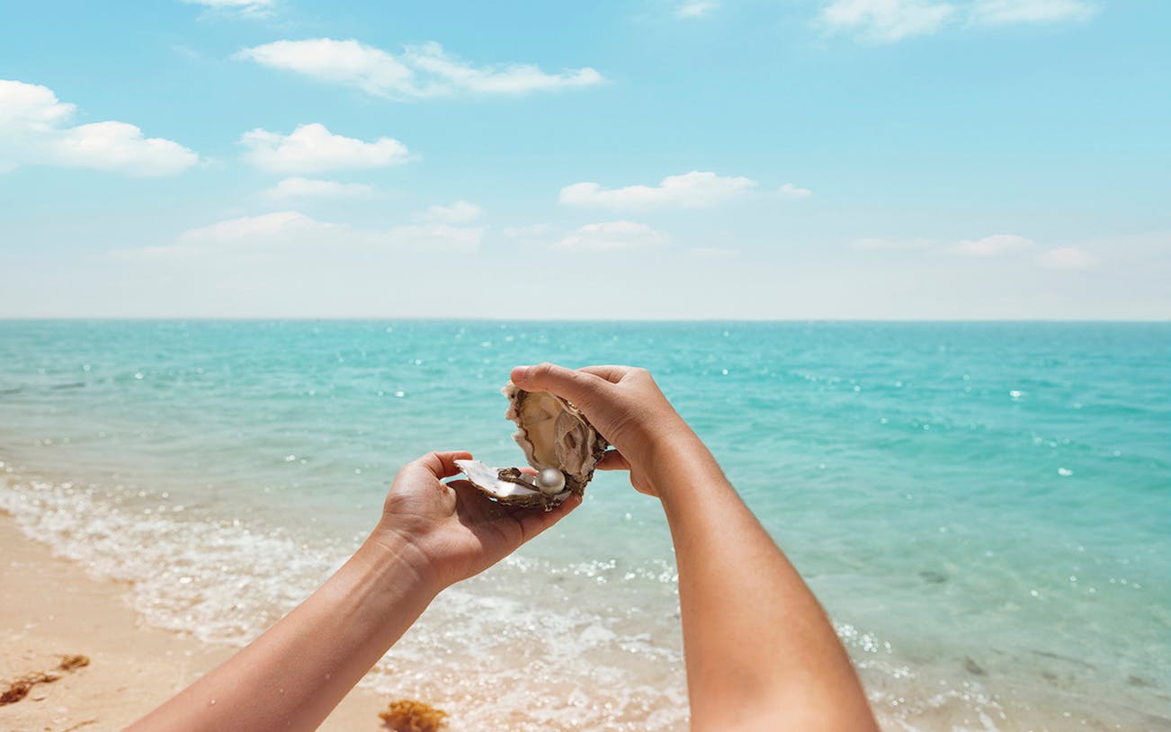 Hands holding an open oyster with a pearl on a beach during Al Suwaidi Pearl Farm Tour.