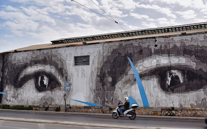 Street art mural of eyes on a building in Athens with a motorcyclist passing by.