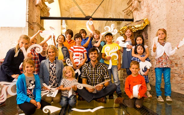 Children and adults holding decorative paper cutouts inside Klosterneuburg Monastery.