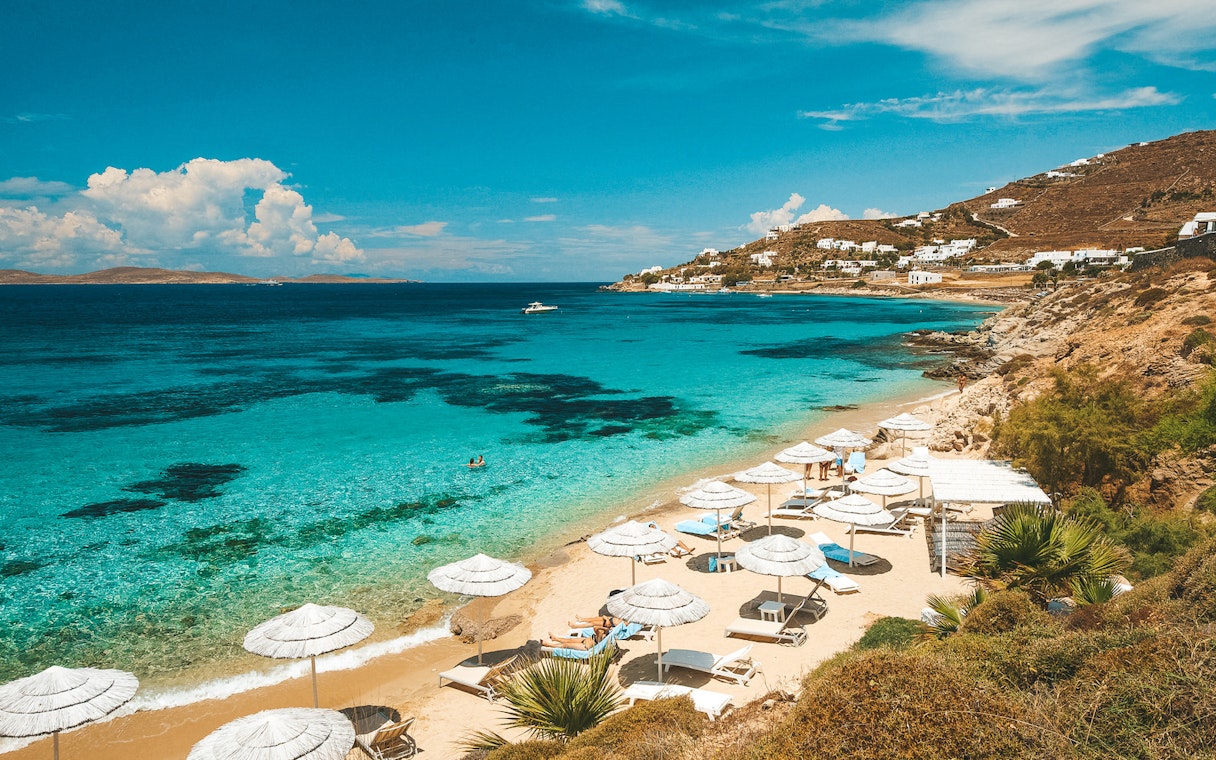 Mykonos beach with umbrellas and loungers, clear blue sea, and distant hills.